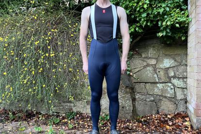 The Rapha Pro Training Tights in navy blue are being worn by a while male who is wearing a sleeveless undervest standing in front of a stone wall with greenery overhanging 