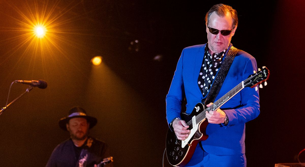 Joe Bonamassa wears a blue suit, polka dot shirt and shades as he takes a solo on his signature Epiphone Les Paul. Josh Smith is in the background on rhythm guitar.