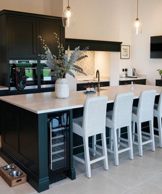 dark green kitchen units, kitchen island with white worktop, white wicker bar stools and glass pendant lights above as well as hob area recessed in lit alcove on back wall