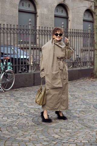 Renia Jaz wears a beige trench coat, black shoes, and a green bag outside during Copenhagen Fashion Week Fall 24.
