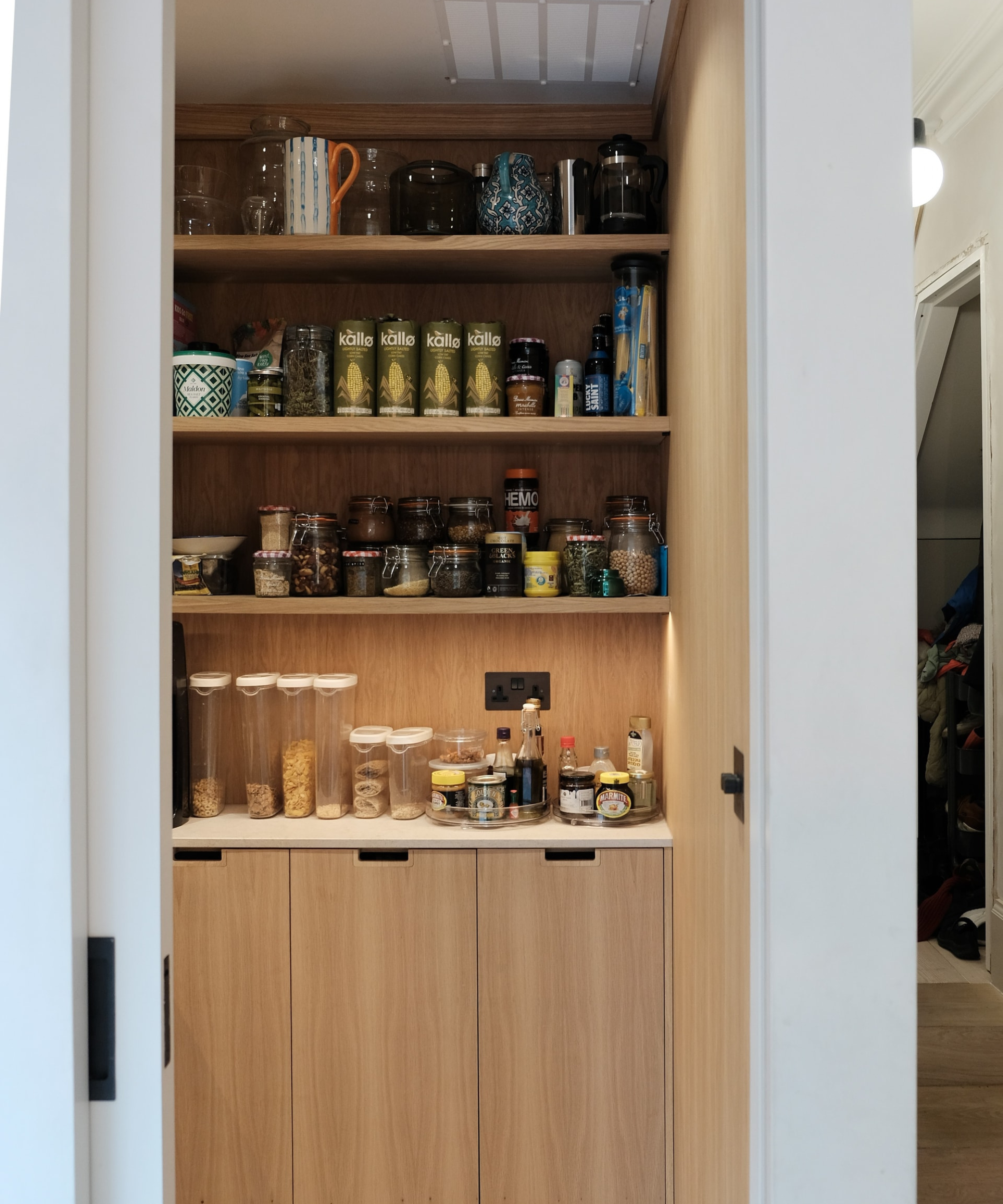 A compact larder cupboard reveals organised shelves filled with jars, dry goods and condiments. Warm wood interiors, integrated lighting and minimalist cabinet doors create a calm, efficient space designed for easy access and clutter-free kitchen storage.