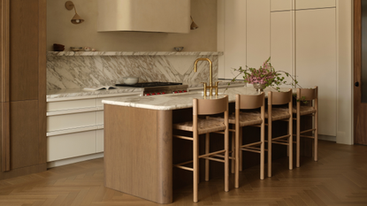 A gorgeous kitchen with a curved concealed exhaust, a marble backsplash, a wood island with a stonetop, and a set of woven stools
