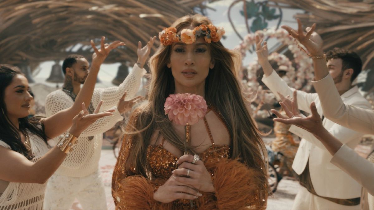 Jennifer Lopez walking down an asile of people with their hands up, she&#039;s wearing an orange dress and flower crown while holding a big pink flower.