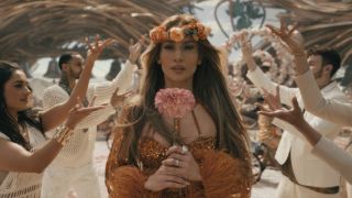 Jennifer Lopez walking down an asile of people with their hands up, she's wearing an orange dress and flower crown while holding a big pink flower.