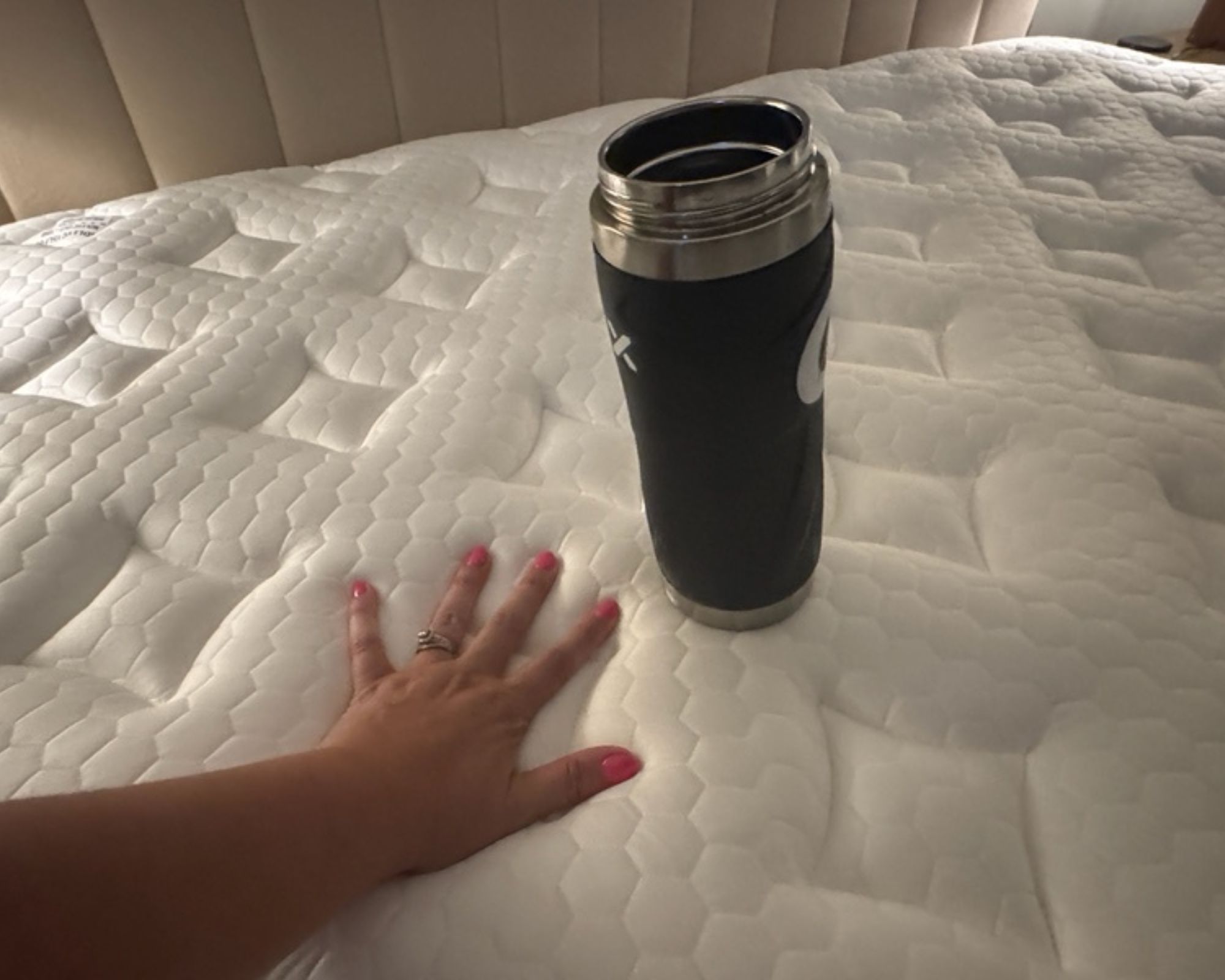 A woman's hand testing the motion isolation of the white Nolah Evolution mattress with pink nail polish, beside a black water flask.