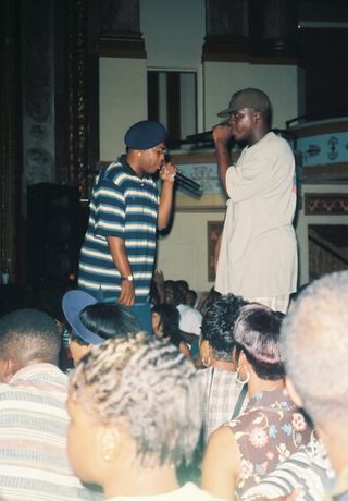 Two young rappers dressed in oversized T-shirts, a stripy white-and-blue one and a gray one with a print on its back, sing in the middle of a crowd in a sweaty night club.