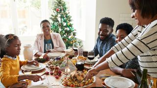 Family gathered round a turkey and sides with glasses of wine on Christmas Day with sparkling tree in the background