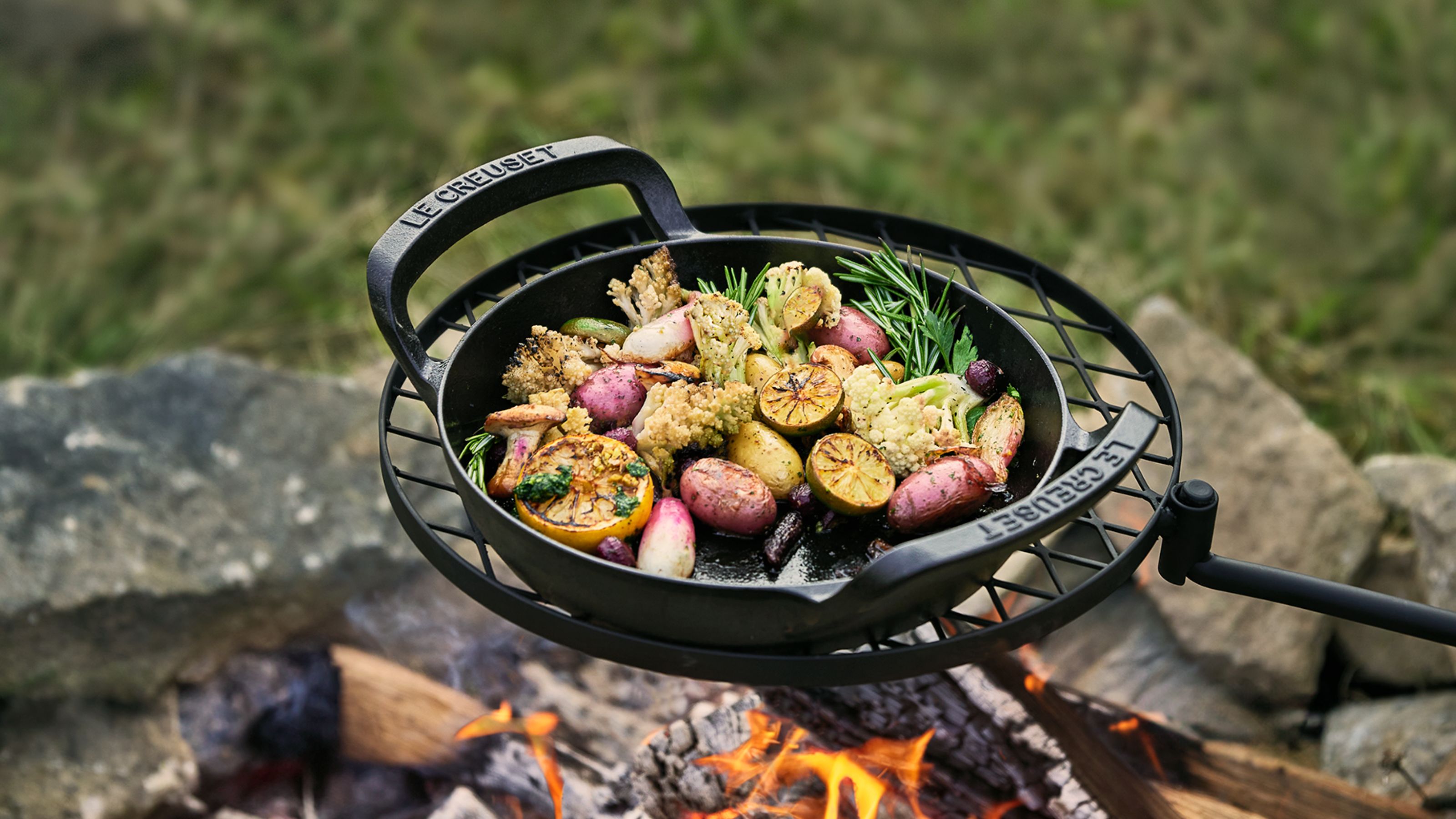 a le creuset cast-iron pan held over an open fire with vegetables cooking in it