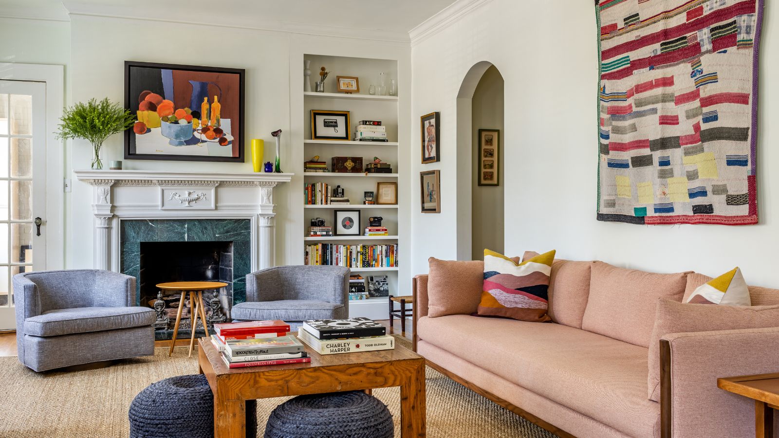 Living room with white walls, arched doorway, pink sofa, jute rug, a wooden table and navy blue pouffes, marble fireplace, and an alcove of books