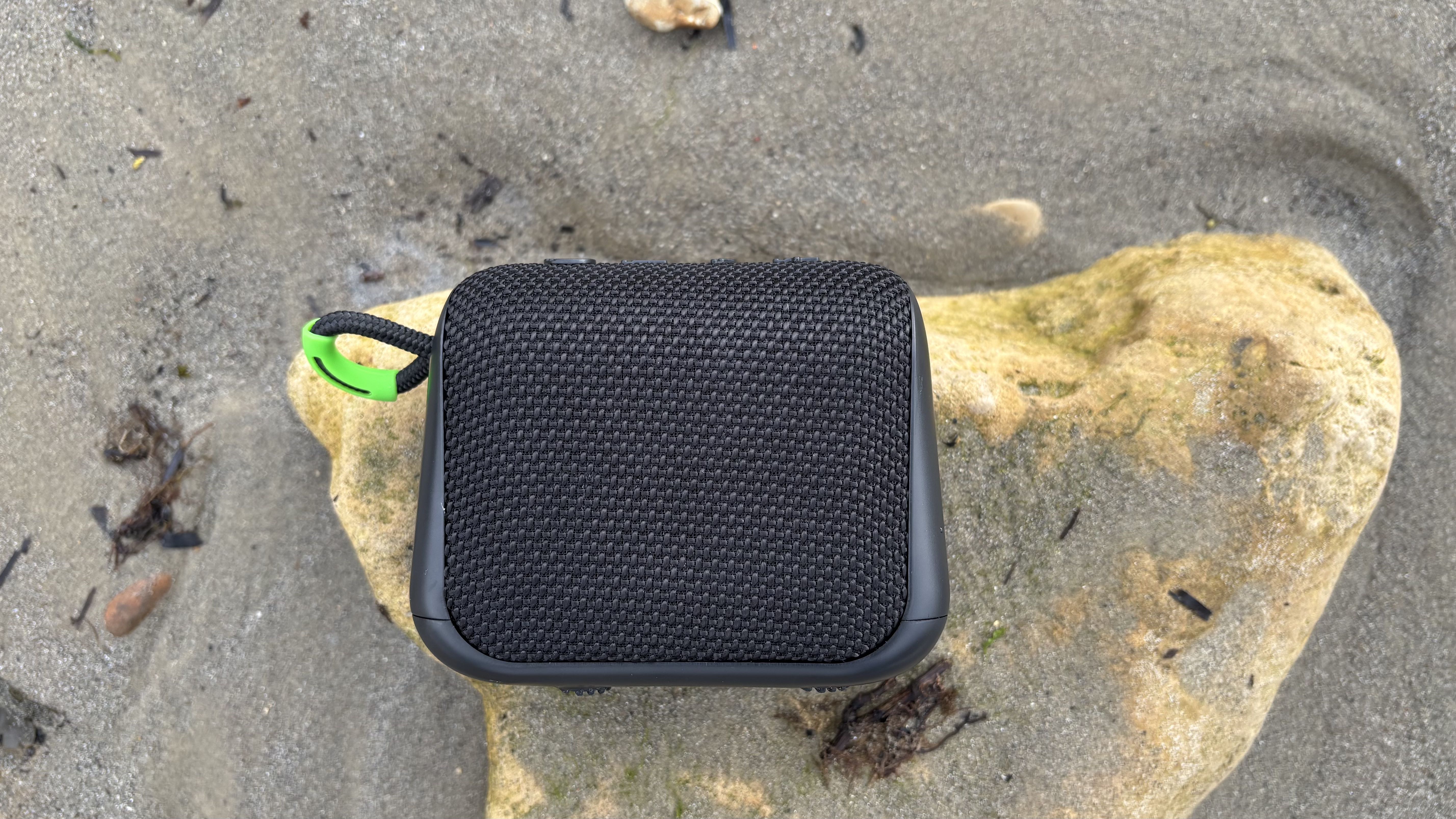 Black AO mini portable wireless speaker on a rock, surrounded by a sandy beach