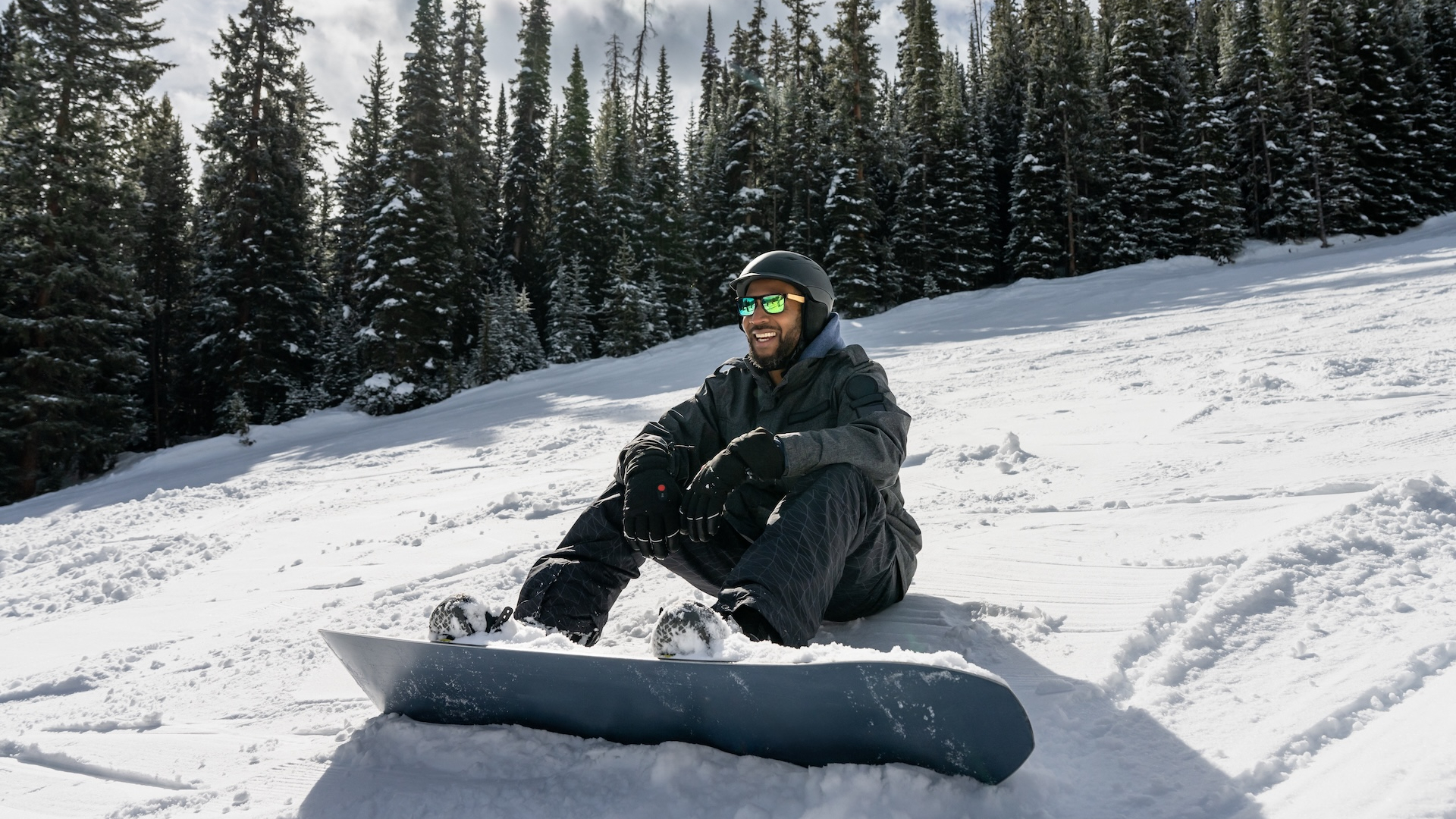 Portrait of a man in his thirties sitting on the snow with his snowboard strapped to his feet at a ski resort in Winter Park, Colorado.