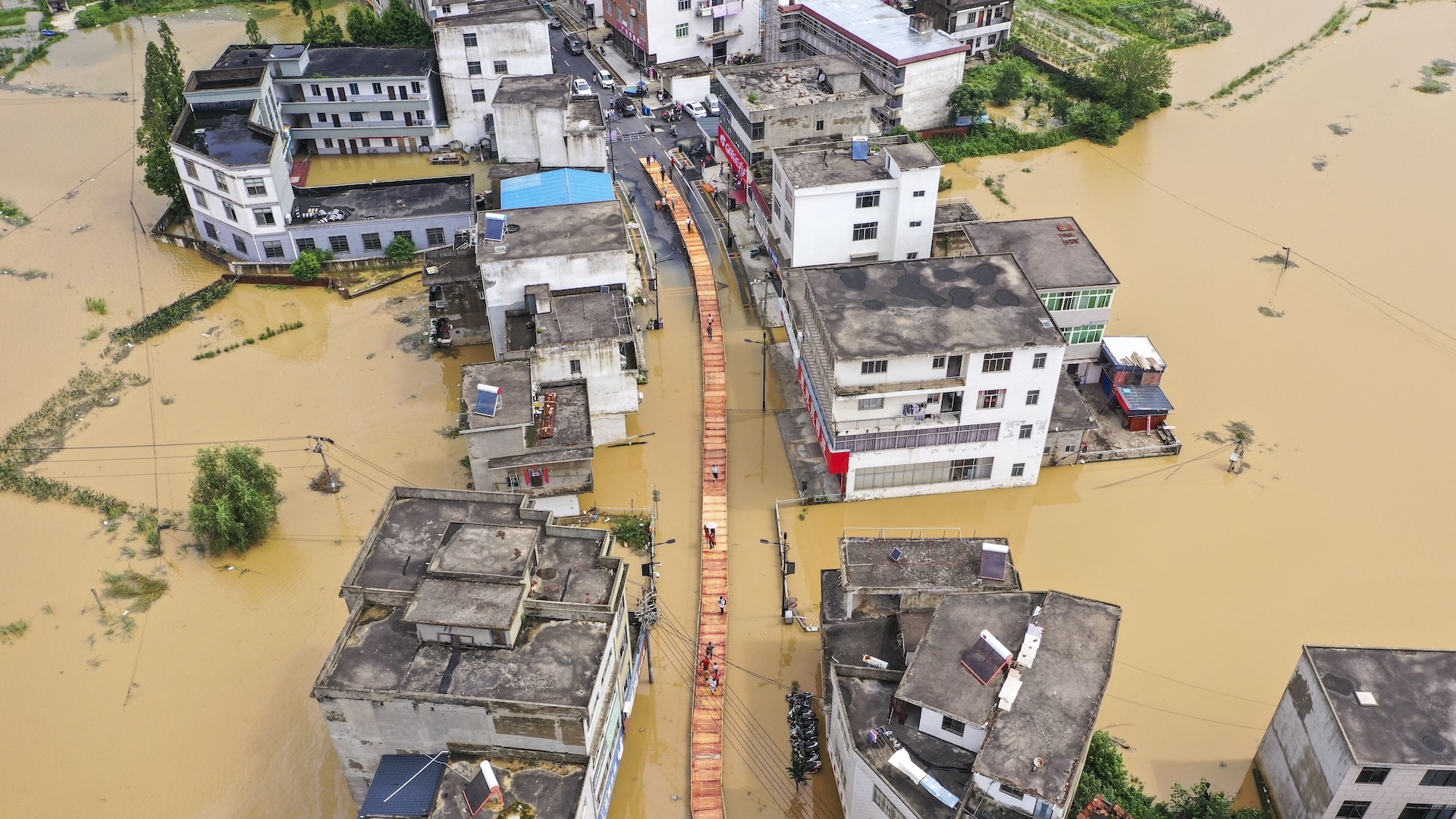 an aerial photograph shows a street of buildings all submerged in water and a central walkway build above the floodwaters in the center, people are walking along it.