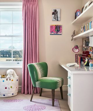 A child's bedroom with beige walls, pink patterned curtains and a green upholstered desk chair.