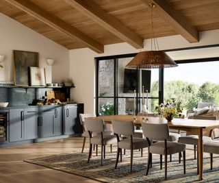 Upholstered chairs around a wooden dining table, topped with candles and a vase of flowers; in the background, dark cabinetry and open French doors looking onto a garden view.