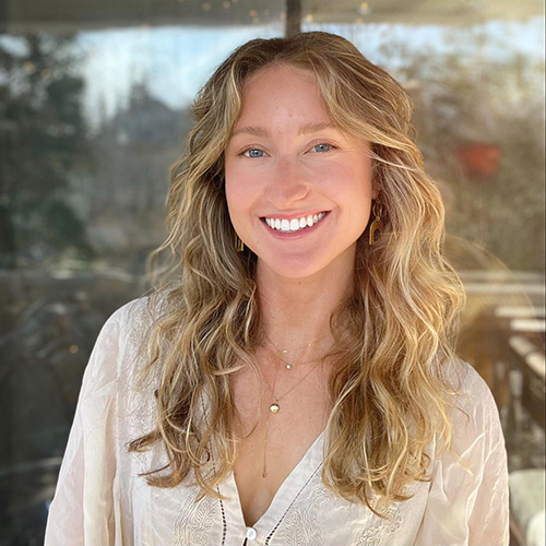 A woman with blond wavy hair wearing a white long-sleeve shirt smiles for the camera.
