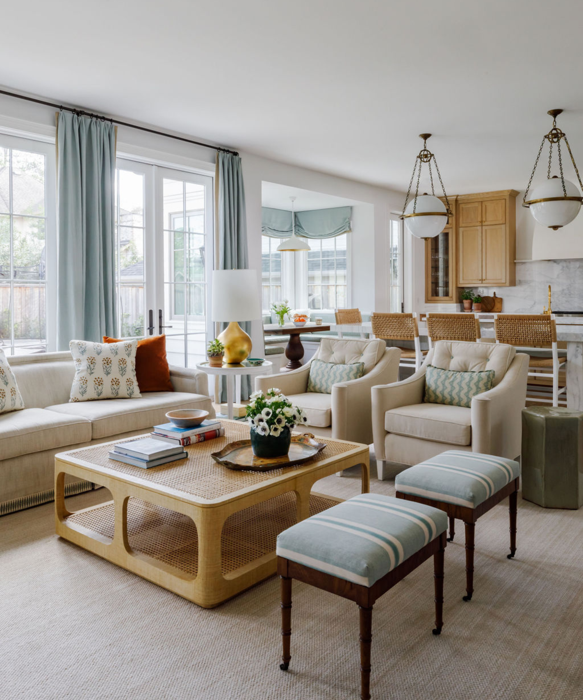 Bright, coastal-inspired living room featuring a large two-tier light wood coffee table, a cream sofa and coordinating armchairs, and blue striped ottomans.