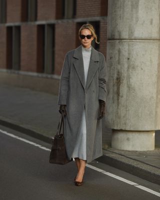 Anouk wears a gray long wool coat, a gray maxi dress, black heels, brown leather gloves, and a brown tote bag.