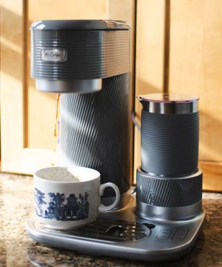 Mr. Coffee 4-in-1 single serve coffee maker in gray colorway in the process of preparing black coffee into a white and blue chinoiserie-style ceramic cup on granite-effect worktop in kitchen with light wooden cabinetry