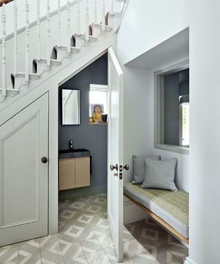 Hallway and staircase with door open to cloakroom, decorated in grey tones with grey and white tiled floor, hallway seat with cushions and