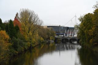 An image showing a river surrounded by trees with a football stadium in the distance