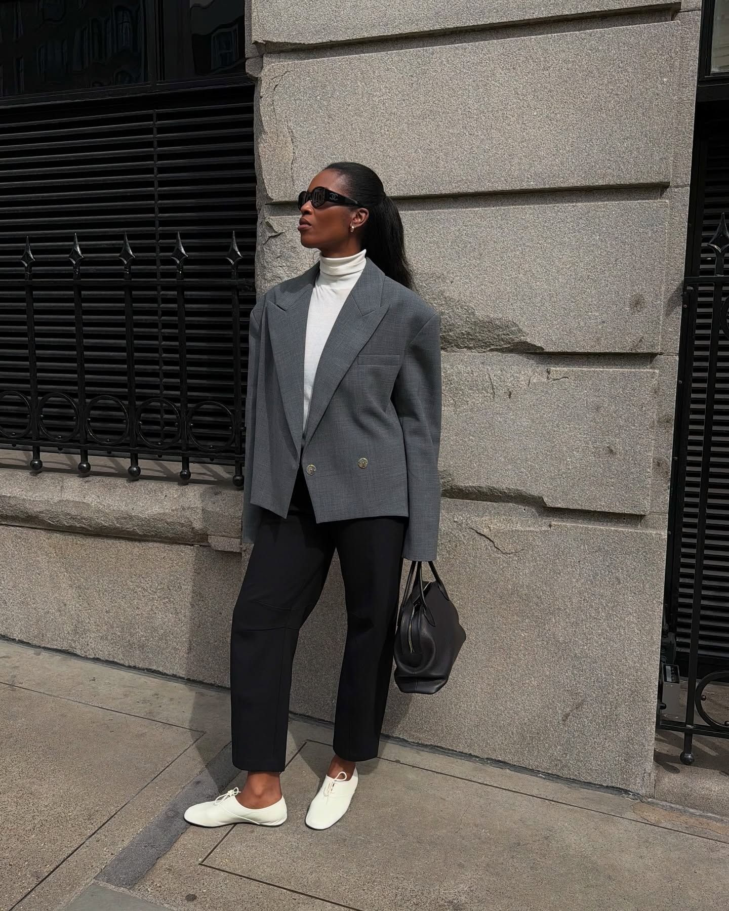 Marilyn wears a gray blazer with a white turtleneck, black trousers, and white derby shoes.