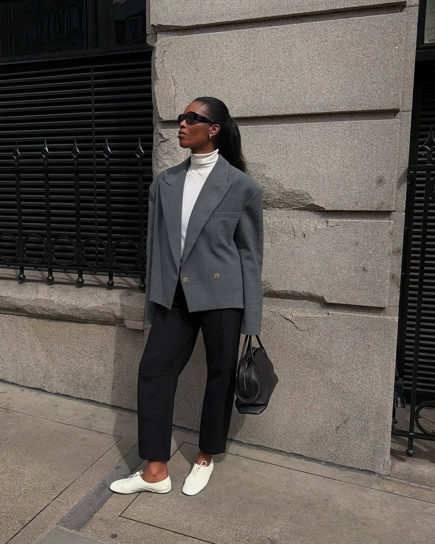 Marilyn wears a gray blazer with a white turtleneck, black trousers, and white derby shoes.