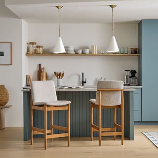 West Elm image of a matt blue-green kitchen island, with two cream upholstered and wooden kitchen bar stools and overhead hanging pendant conical white lamps