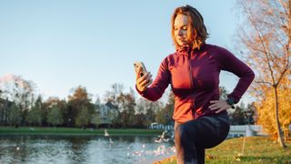 Woman using a workout app on phone, lunging forwards, working out in the park on a cold morning