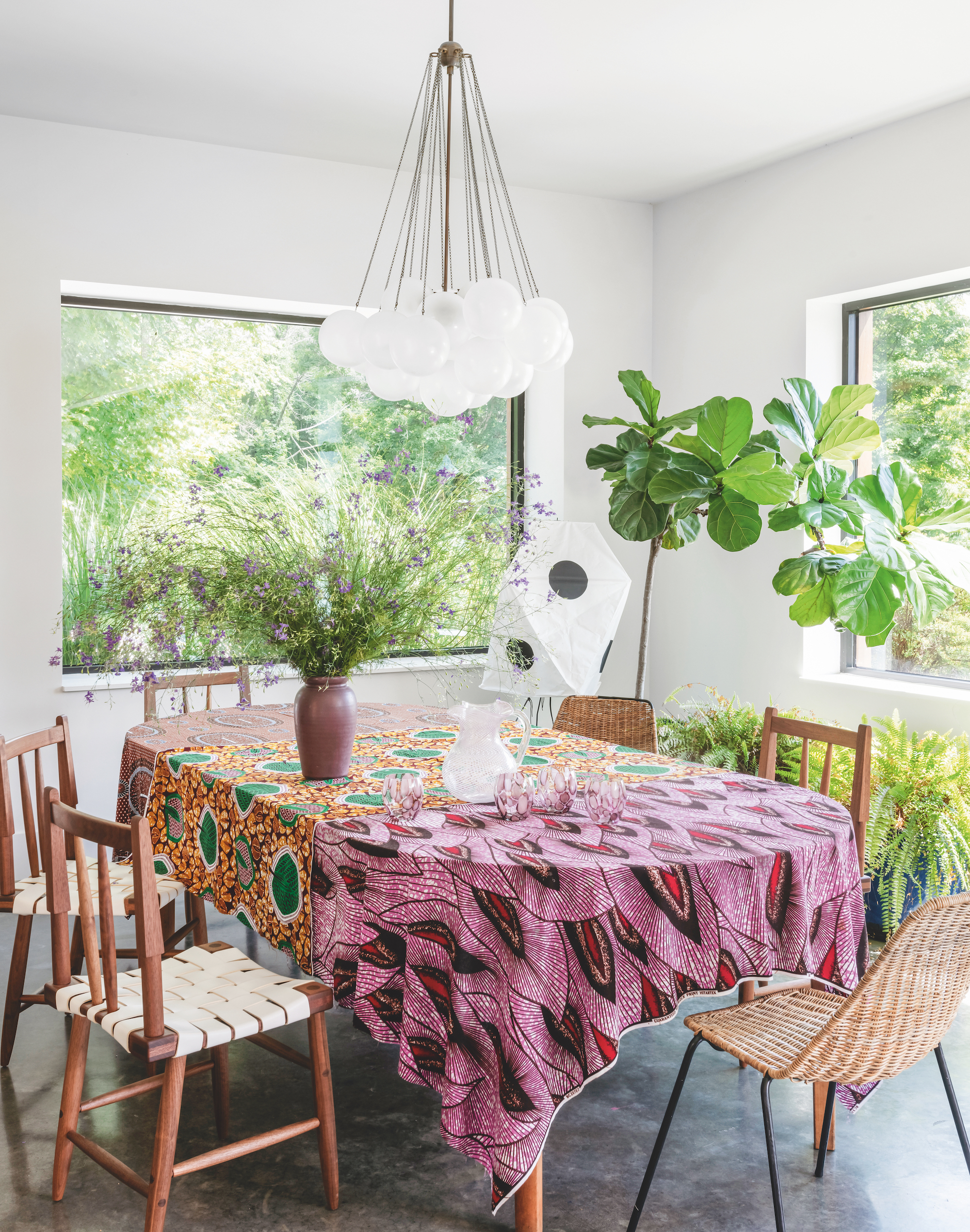 A white walled dining area, with a curved dining table, covered in a pattered table cloth and miss-matched wooden chairs around. Behind the table are some potted plants and there is a modern chandelier hanging above the table.