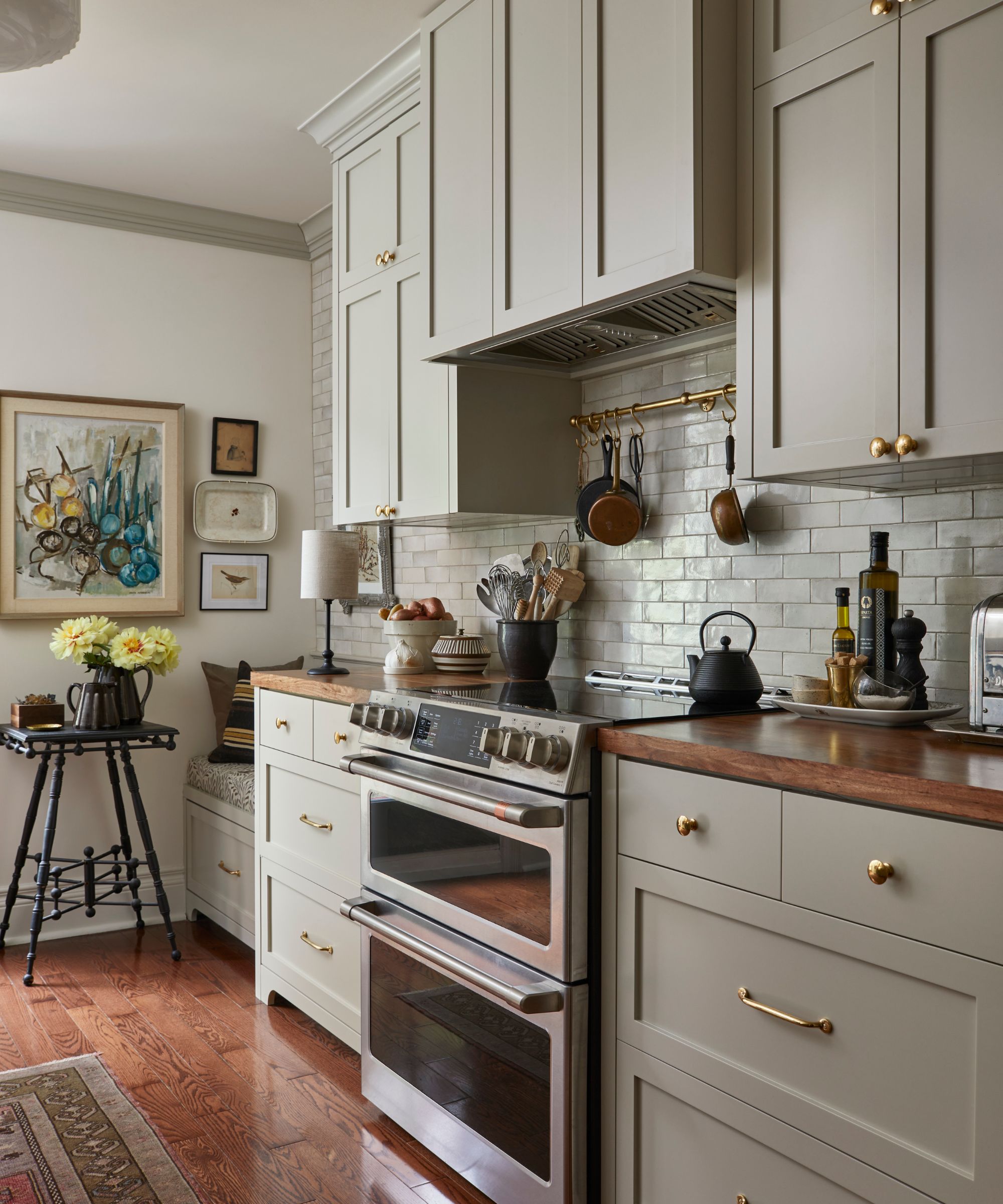 A vintage-inspired galley kitchen with gray cabinets, wood counters, a steel oven, a brass pot hanger, and banquette seating in the corner