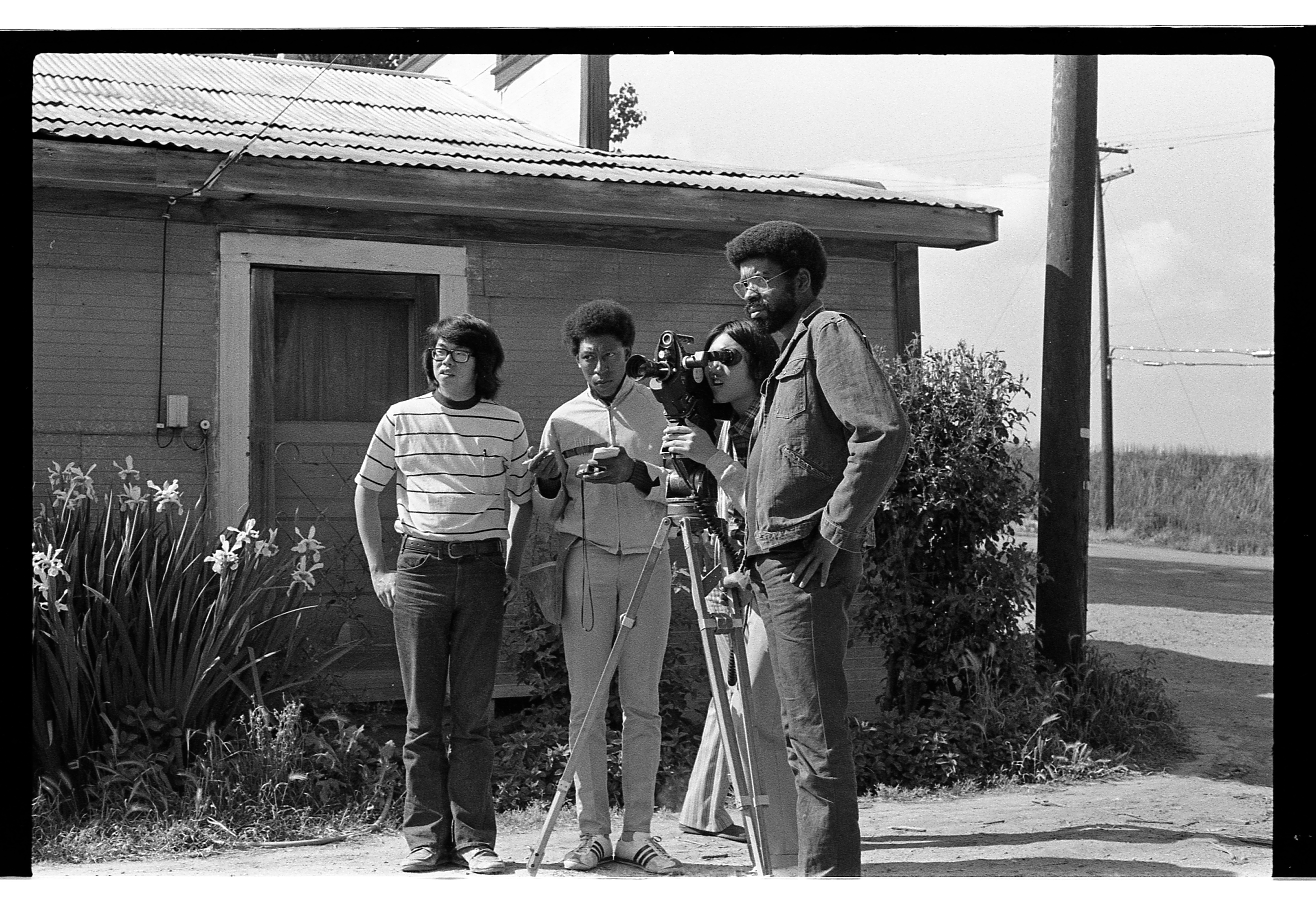 Four people outside a rustic building, setting up and operating a vintage film camera on a tripod in a rural area.