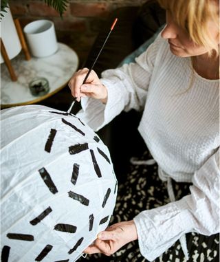 woman painting a white lantern lamp with black stripes, next to a marble side table