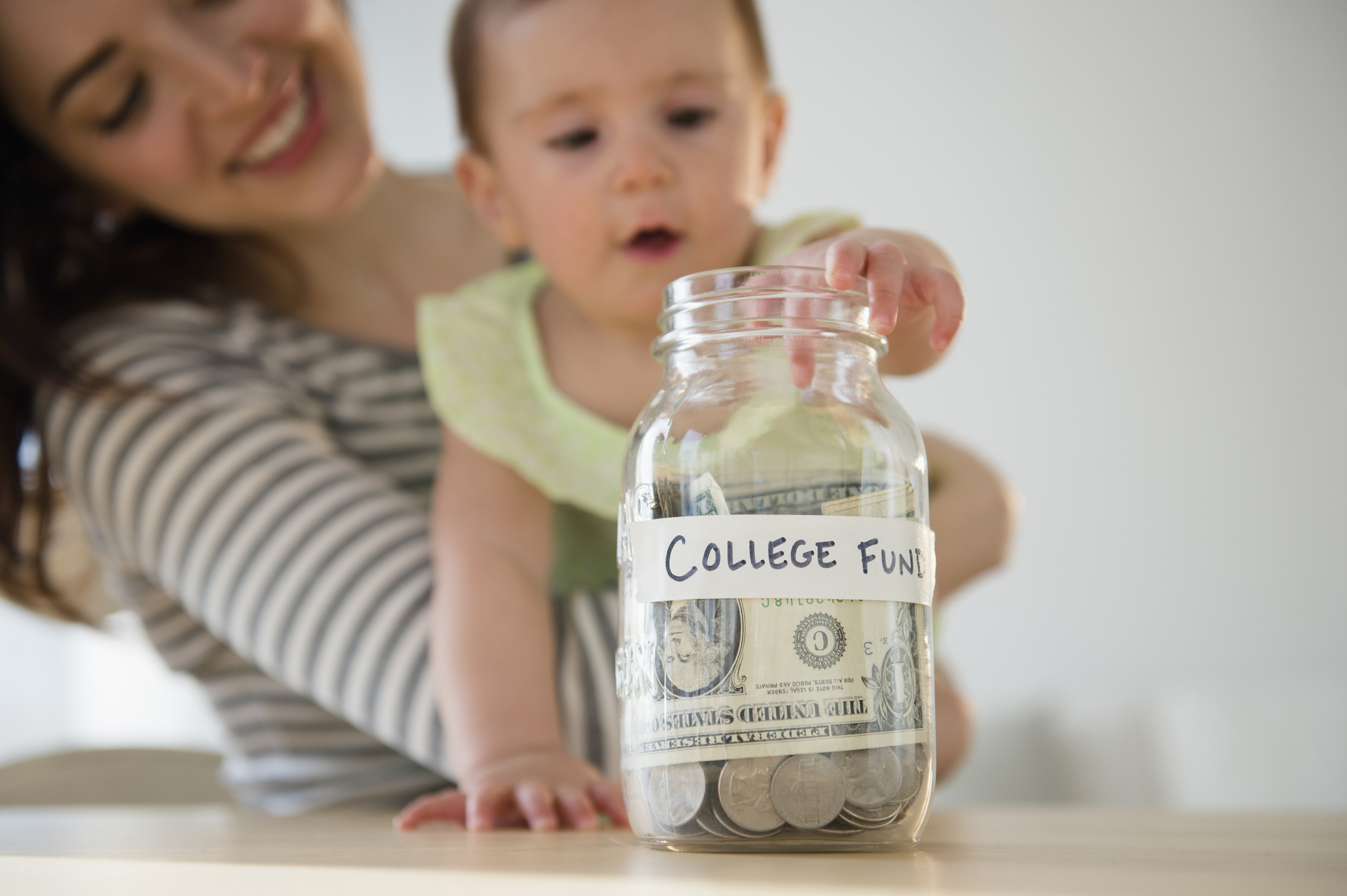 A baby held by her mom deposits a dollar bill into a jar marked college fund
