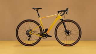 A yellow and orange gravel bike with front suspension stands in front of a yellow background
