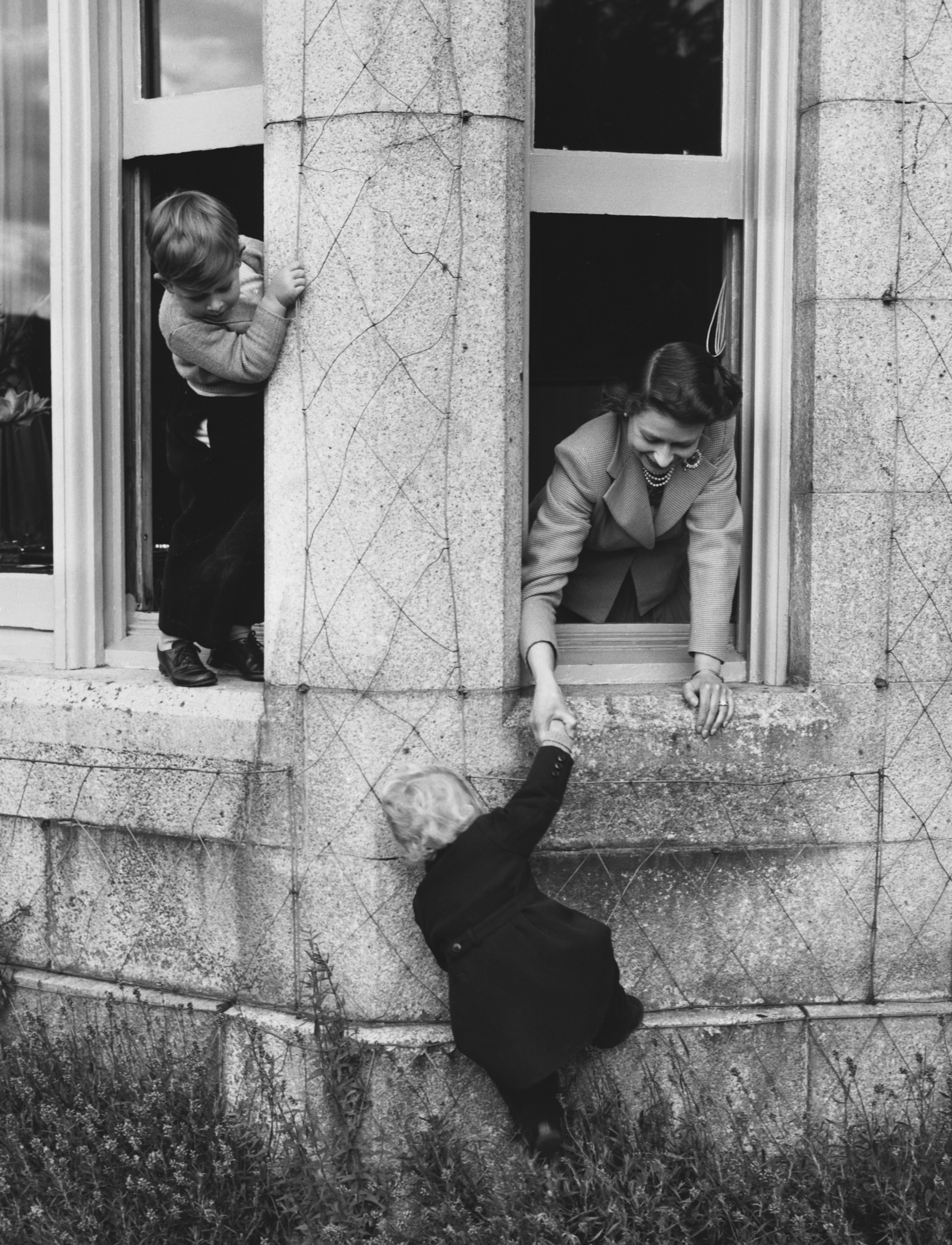 Princess Anne hanging out of a window holding Queen Elizabeth's hand while Prince Charles looks out another window