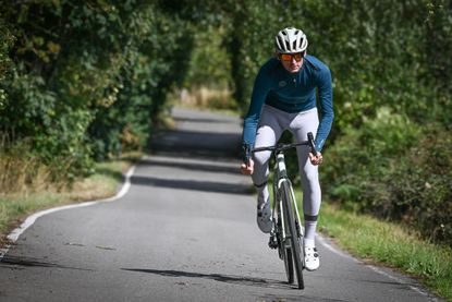 Man wearing a green long sleeved jersey, pale grey tights and a beige helmet riding towards the camera on a country lane