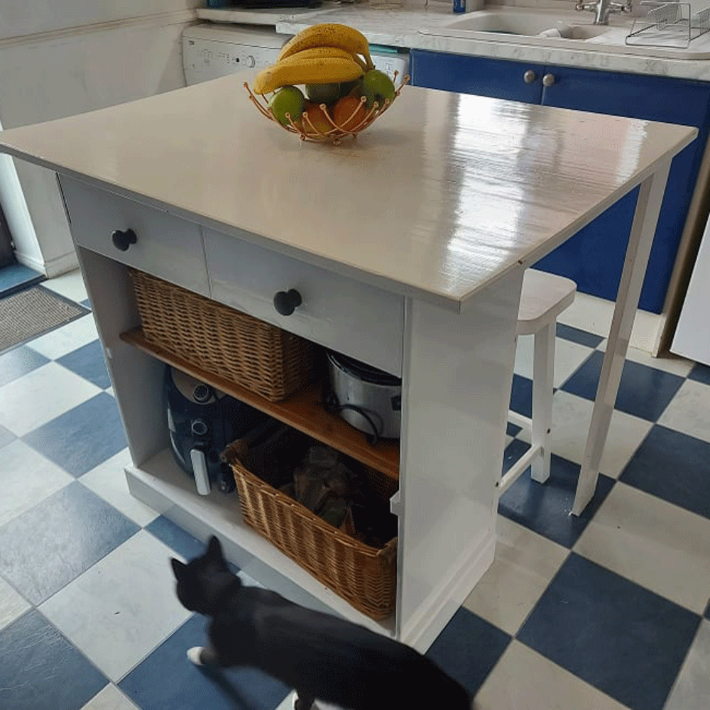 Black and white cat walking past a white kitchen island with 2 drawers and a shelf containing 2 woven baskets, an instant pot and airfryer