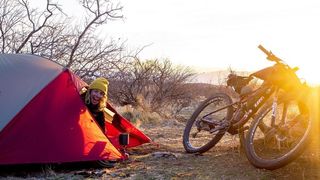 Sarah Sturm pokes her head out of her tent while her bike leans against a rock to the right
