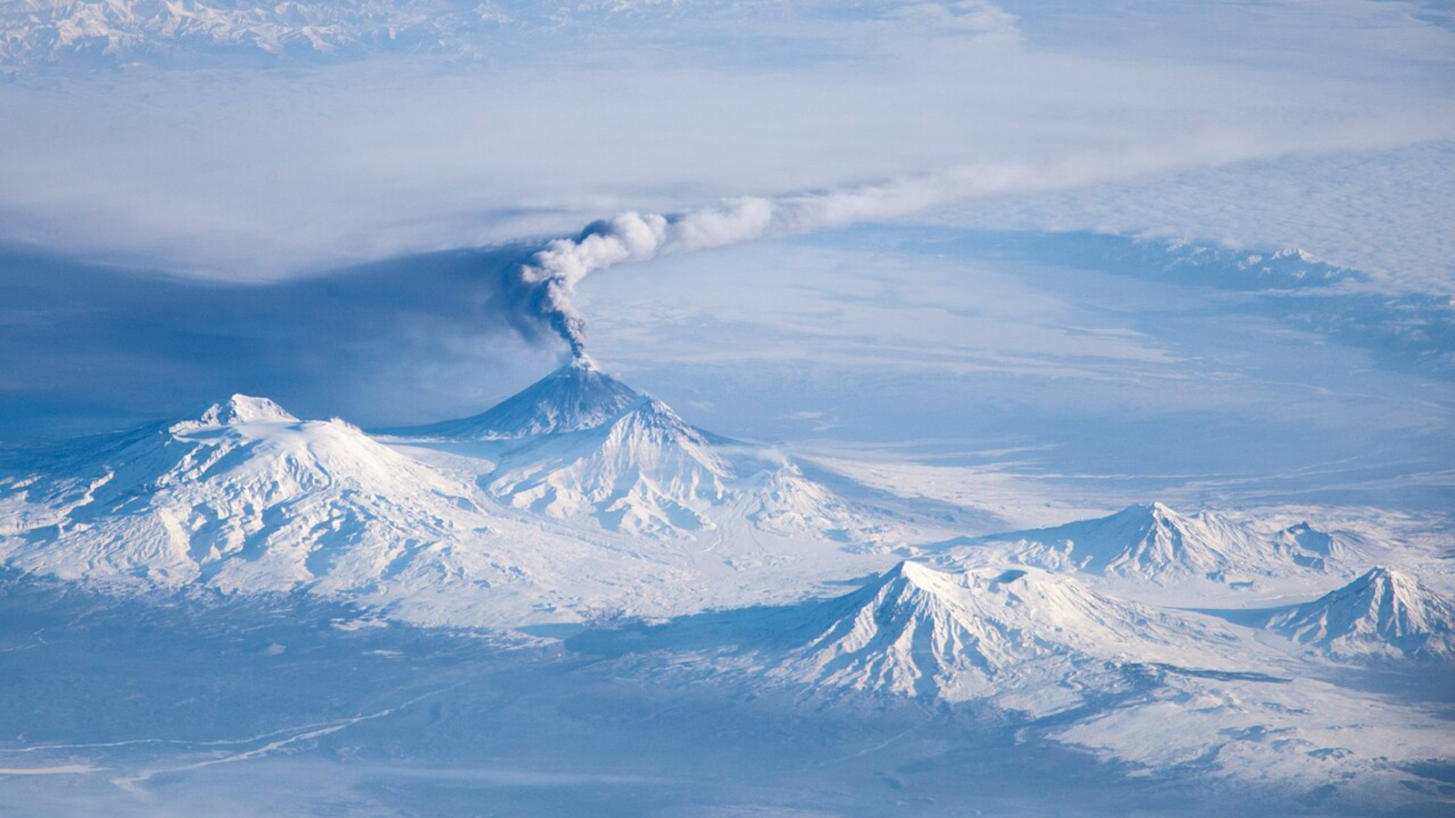 A photo of multiple snow covered volcanoes with an eruption plume coming from one of the peaks