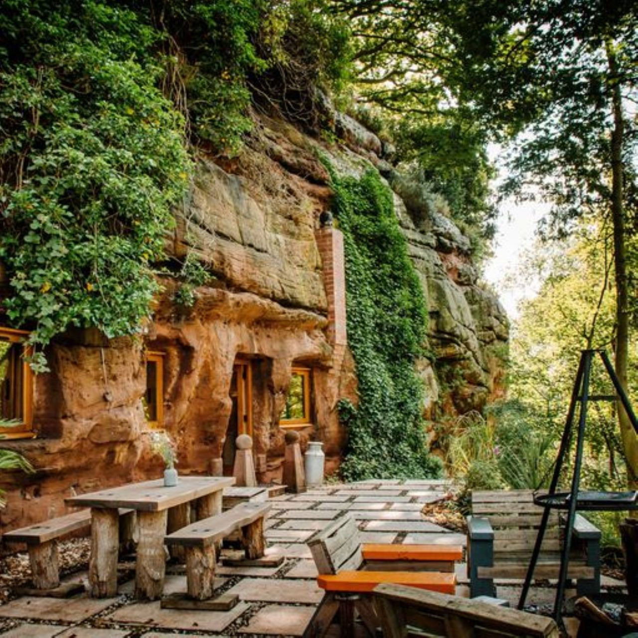 The Rockhouse Retreat entrance shows the side of a rock wall with doors and windows installed into it with a patio and walkway entrance as well as vegetation growing from the walls