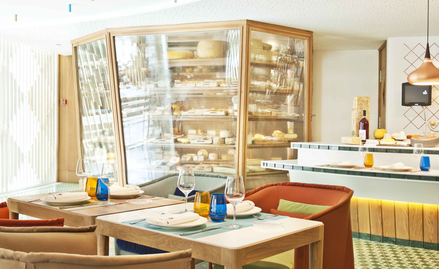 A restaurant with decorated tables, chairs, a pendant light, a food serving counter and a wood and glass cabinet with baked goods inside.