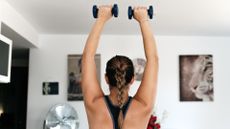 A woman doing an overhead press in her living room with small dumbbells