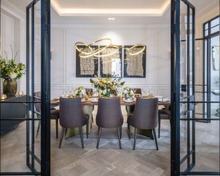A dining room wall idea with crittal doors, grey upholstered chairs, artwork on a white paneled wall and a contemporary gold ring chandelier
