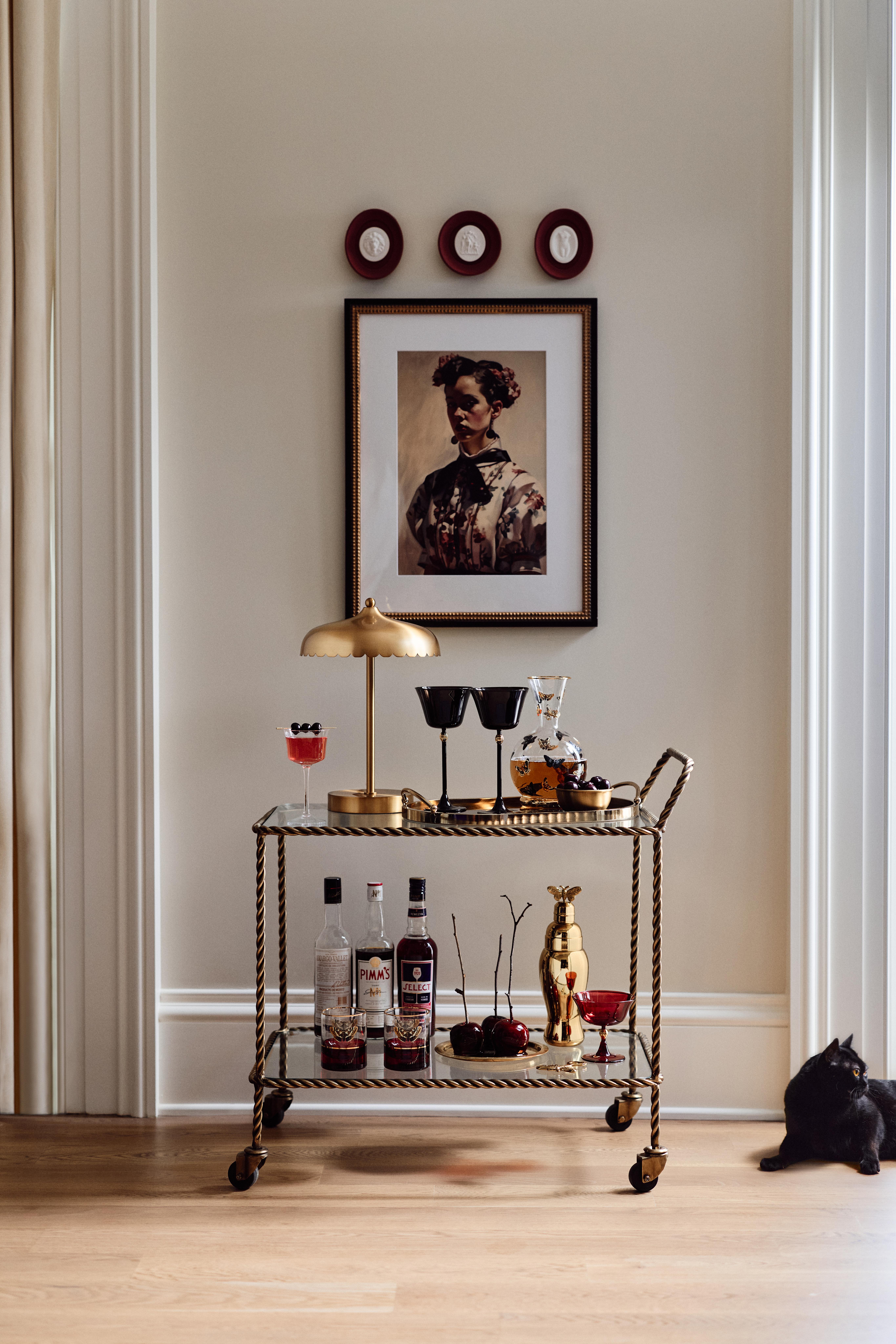 Anthropologie x Catherine Martin Halloween collection barwareon a bar cart underneath a festive framed portrait. To the right sits a black cat