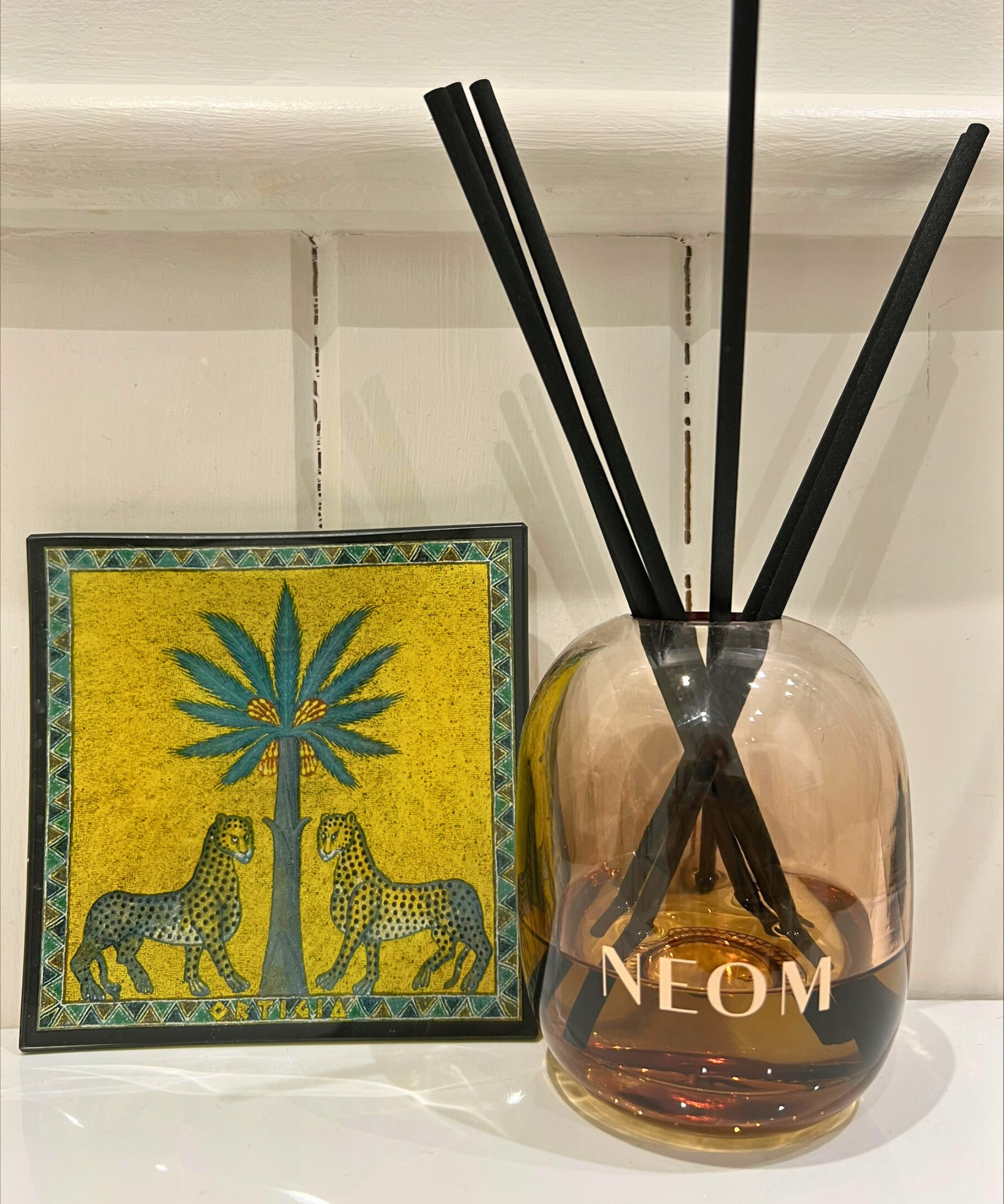 NEOM diffuser beside a yellow Ortigia plate with a white panelled wall behind.