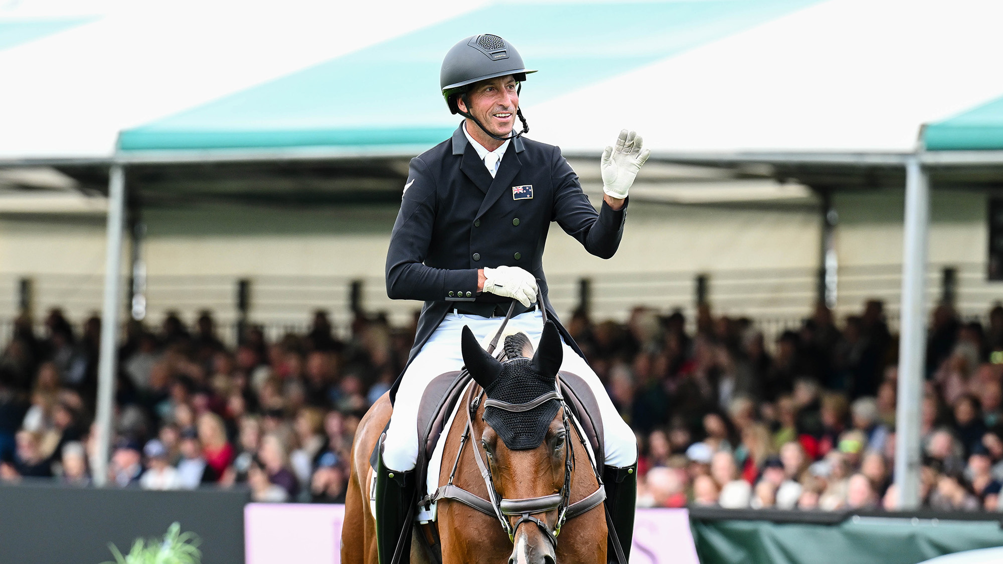 Tim Price, a rider in a black tailcoat, gives a wave as he finishes his dressage test on Falco, a bay horse, at Burghley 2026