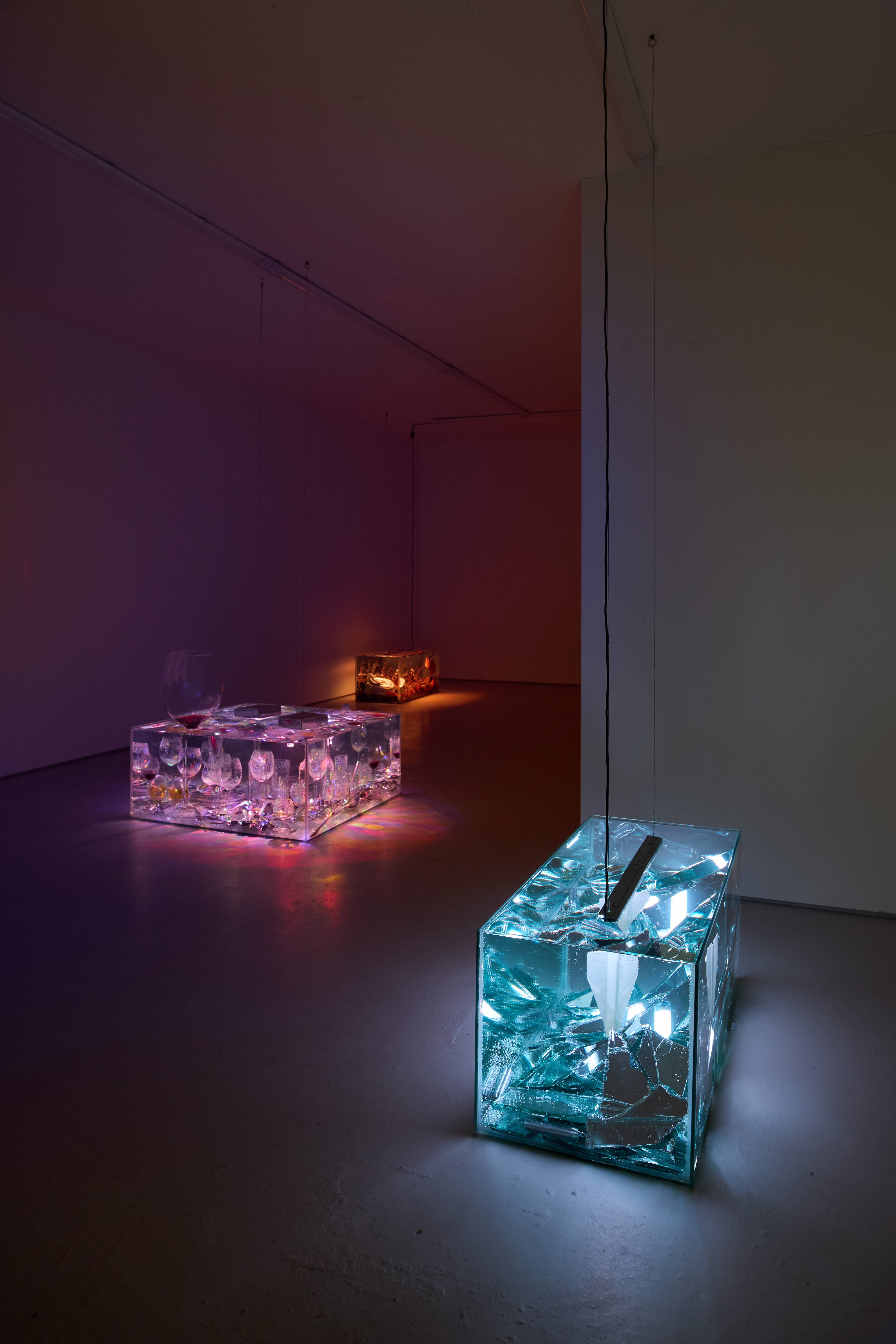 A series of softly glowing, see-through boxes filled with everyday objects like glasses, radios, and domestic tools, unleash warm orange, pink, and pale blue light in a gallery room.