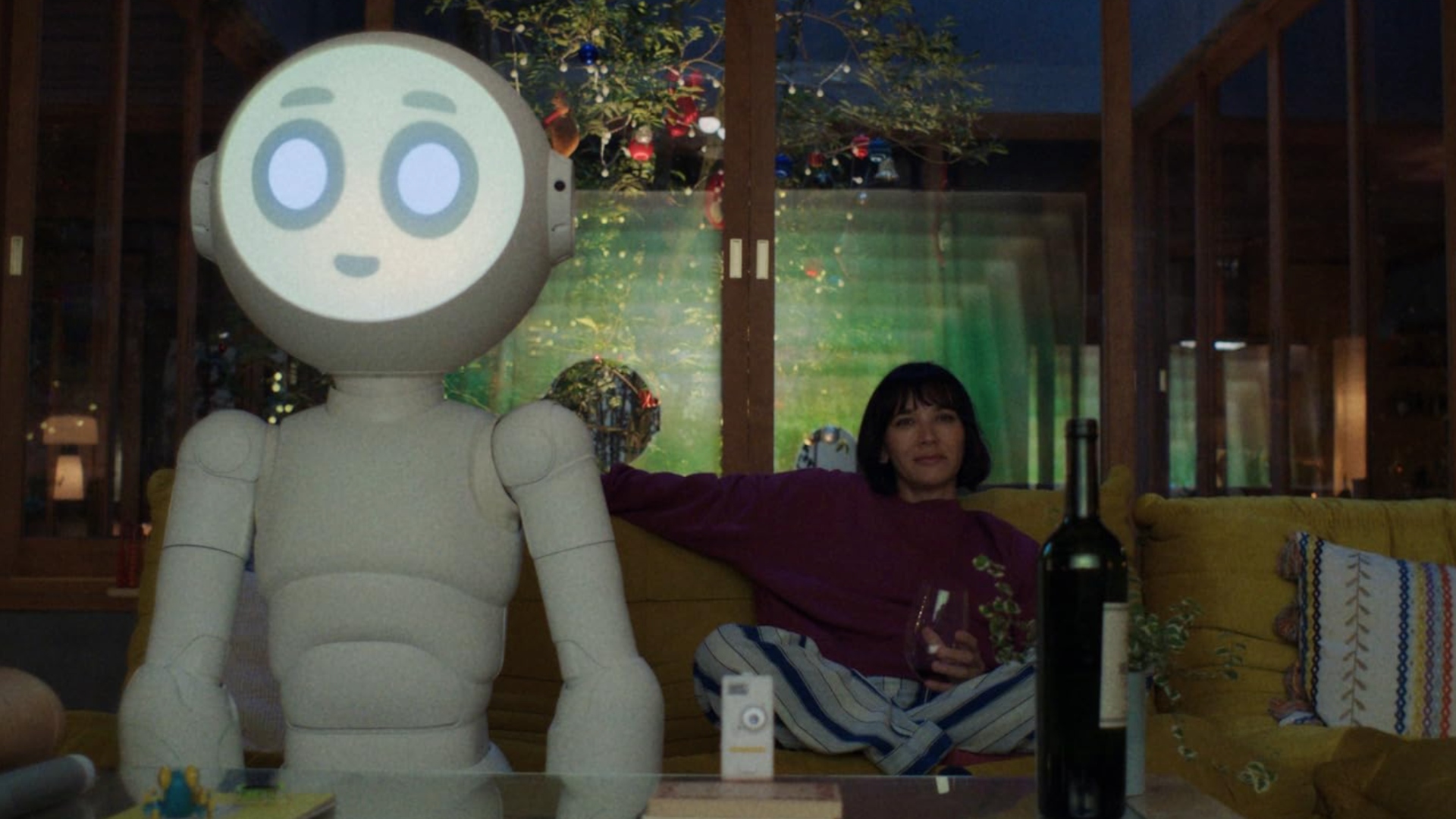 A woman is sitting on a yellow sofa with a bottle of wine infront of her, sitting next to a white robot that has an emoji for a face.