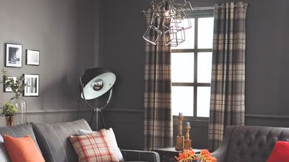 A grey living room with checked eyelet curtains on the window and pops of orange through cushions and flowers