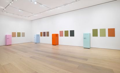 Art gallery featuring white walls, while ceilings and brown laminated flooring. On display are 5 colourful fridges (pink, light blue, orange and light green) spread across the room against the white wall with plain coloured wall art on the walls
